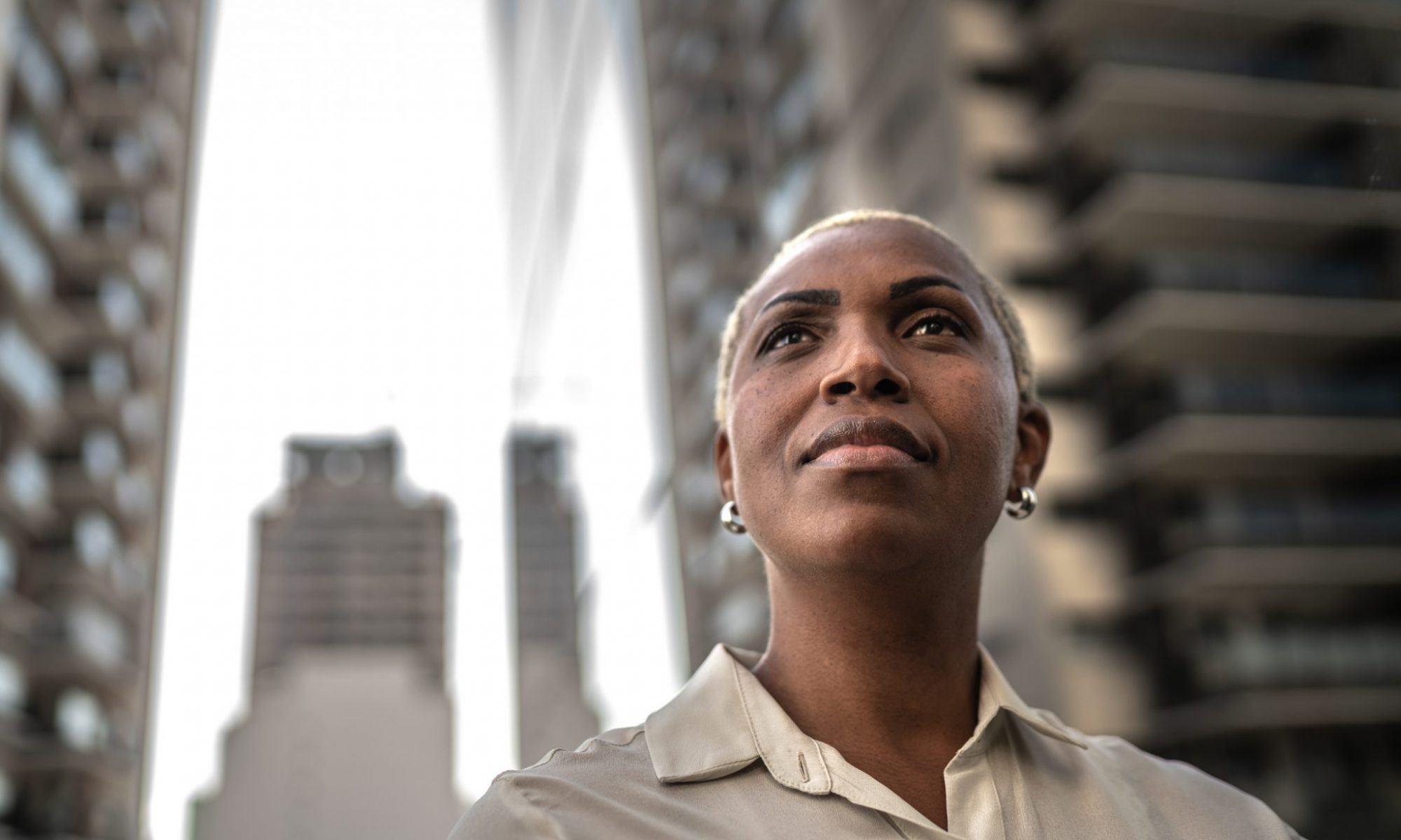 Here is a person with short blonde hair and dark skin wearing small hooped earing looking upwards with curiosity. This individual is wearing a silky sand coloured shirt with an unbuttoned collar.