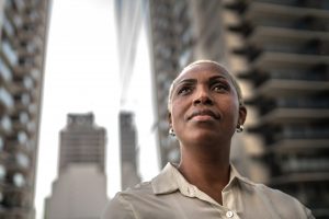 Here is a person with short blonde hair and dark skin wearing small hooped earing looking upwards with curiosity. This individual is wearing a silky sand coloured shirt with an unbuttoned collar.