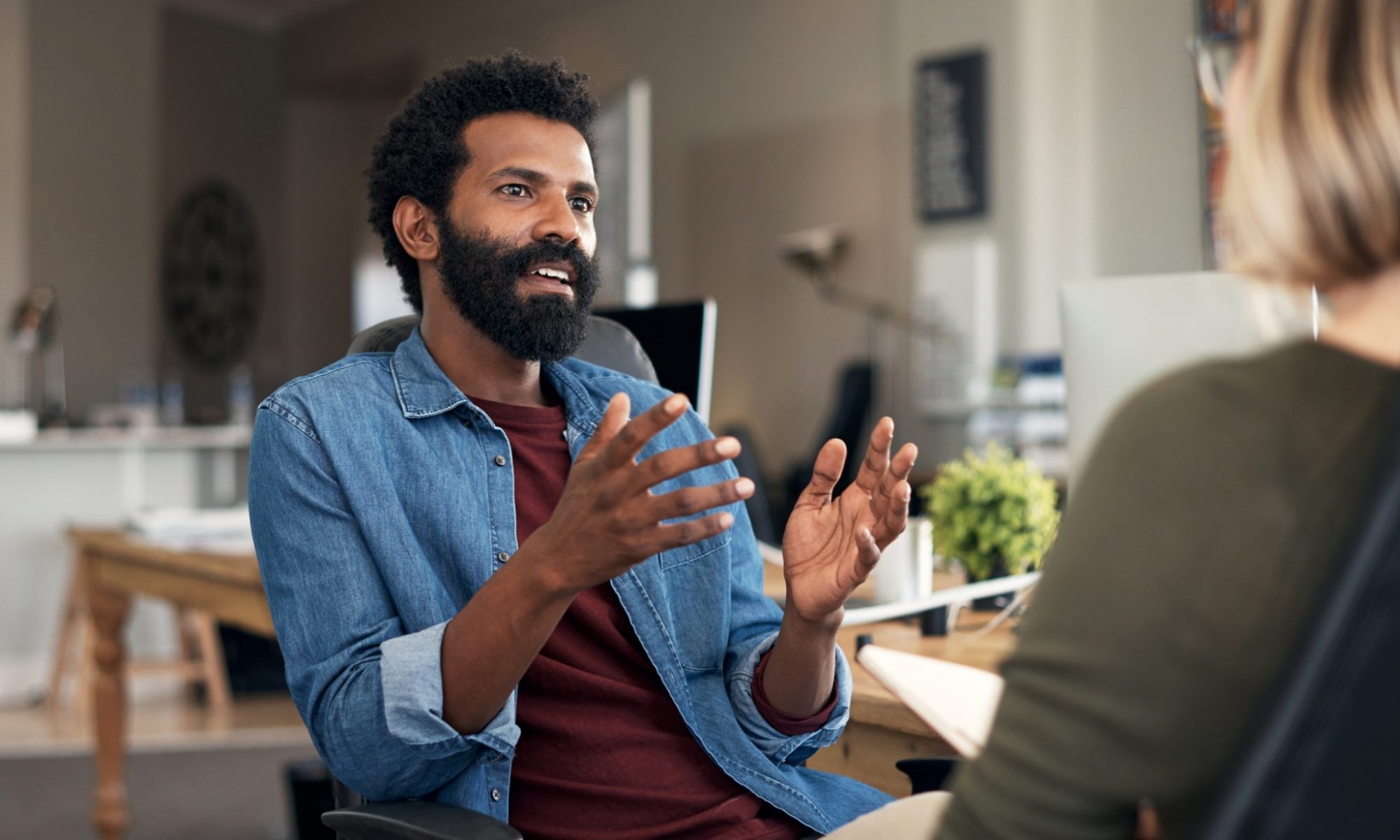 Here are two casually dressed individuals in a closed meeting in a creative office environment. The individual has short black afro hair with a black beard dressed in a maroon tee shirt and a denim long-sleeved on top.
