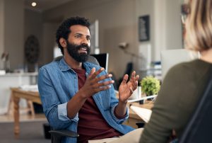 Here are two casually dressed individuals in a closed meeting in a creative office environment. The individual has short black afro hair with a black beard dressed in a maroon tee shirt and a denim long-sleeved on top.