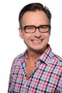 The independent LGBT+ Researcher at the Technical University of Munich and BGA webinar speaker is grinning. The Researcher has brown hair and hazel eyes and wears thick black eyewear frames. Also wears an unbuttoned collar, blue and pink checked shirt.