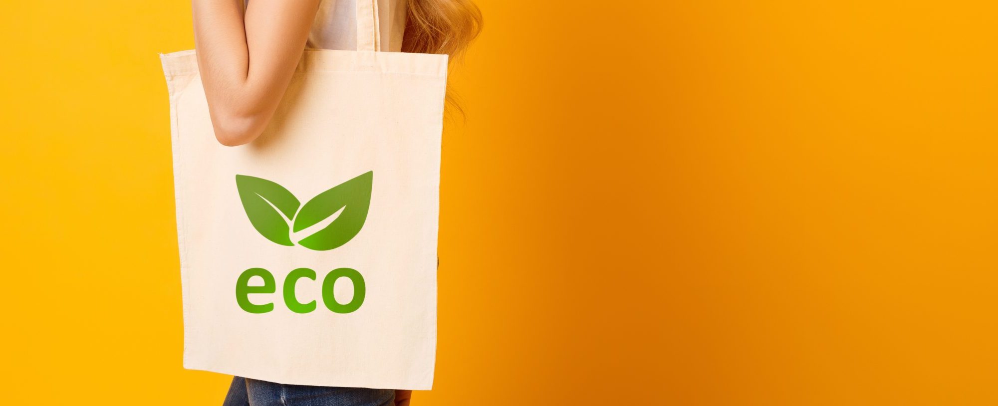 A person is carrying an eco-friendly fabric shopping bag with a green leaf logo stating 'eco'. The person is standing behind a bright solid orange background.