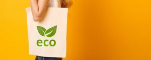 A person is carrying an eco-friendly fabric shopping bag with a green leaf logo stating 'eco'. The person is standing behind a bright solid orange background.