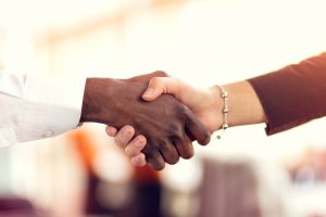 Two diverse hands are shaking. One hand has a long white sleeve, and the other has a silver bracelet with a black shirt sleeve—Symoblic to partnerships and diversity.