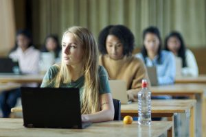 Students are taking an assessment, sitting on individual wooden desks with laptops on top. A person in a green top is sitting in deep thought. The student leans on the table with a satsuma and a water bottle next to the elbow.