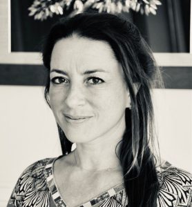 Black and white photograph of the Dean, Mediterranean School of Business (MSB) grinning. The Dean has long straight hair, half up and half down, wearing a thin chain around the neck and wearing a V neck patterned top.