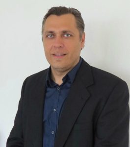 A person with greyish blue eyes with hair slicked back. He has dressed in a dark navy blue shirt with the collar button undone, with a smart black blazer.