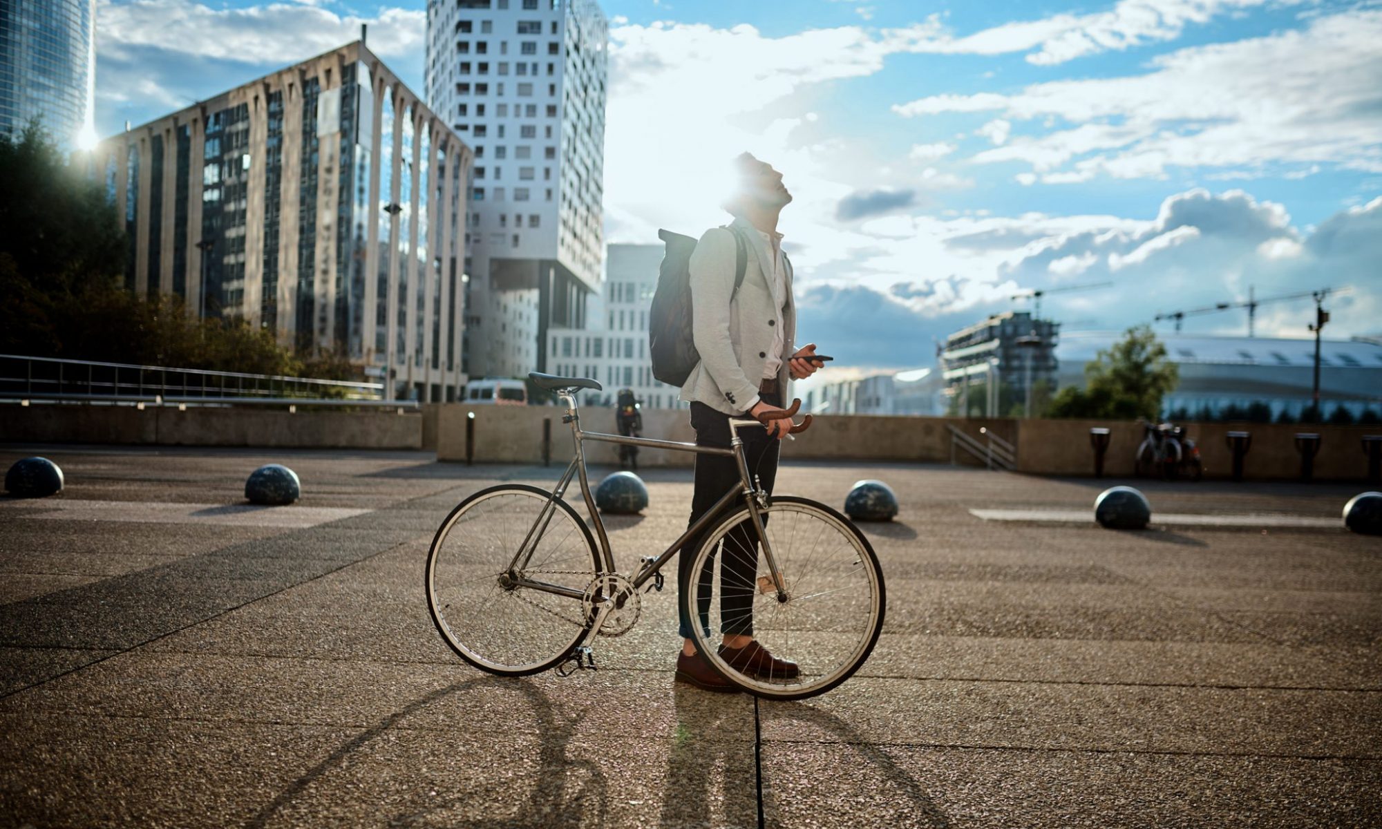 A student is looking into the bright blue sunny sky with a backpack on his shoulders, holding a mobile in one hand and resting the other on a bike. The student is in the city with high glassed buildings.