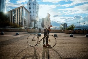 A student is looking into the bright blue sunny sky with a backpack on his shoulders, holding a mobile in one hand and resting the other on a bike. The student is in the city with high glassed buildings.