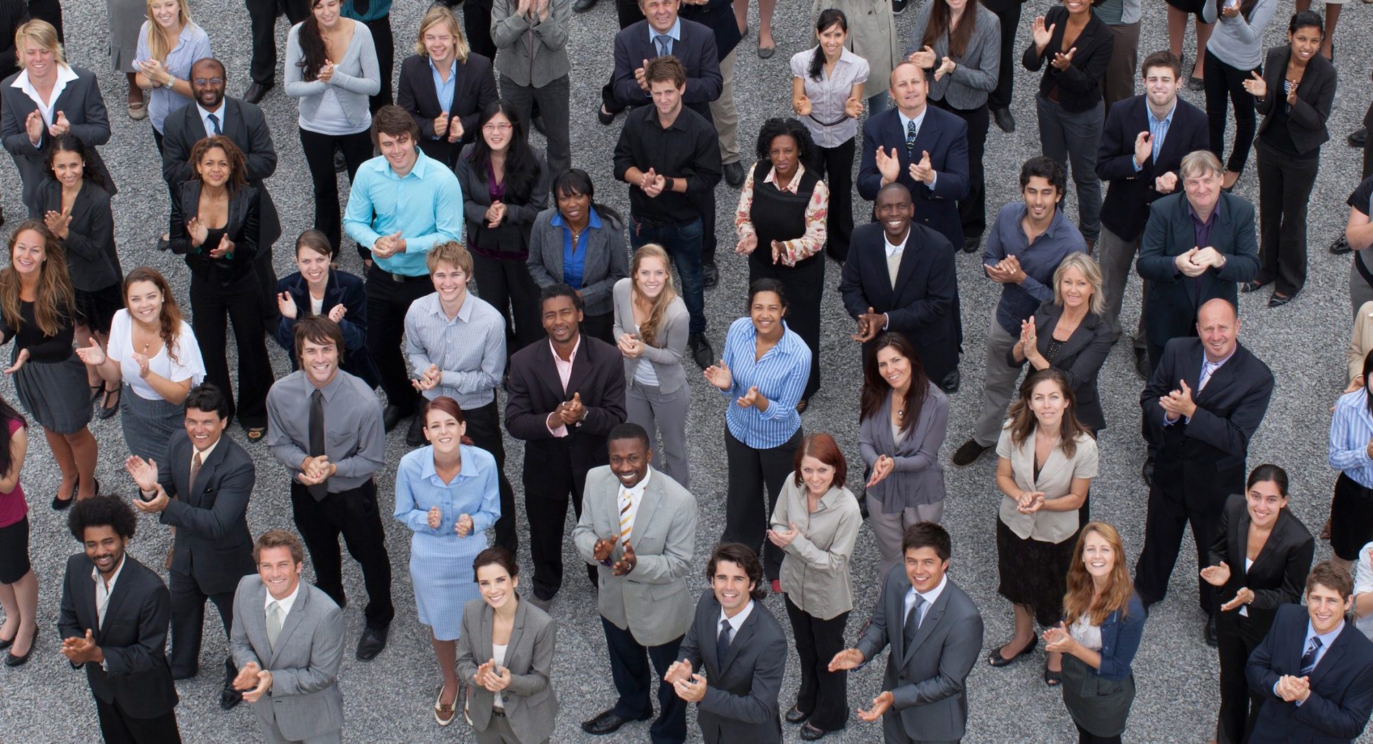 Here is a large, diverse crowd of business working professionals socially distancing standing outside dressed in smart attire.