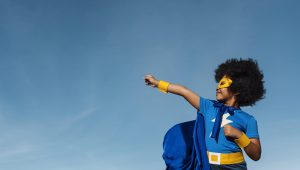 Here is a kid with short black afro hair punching into the sky. The young kid is in a blue and gold superhero outfit with a mask, cap and a lightning bolt icon on his t-shirt.