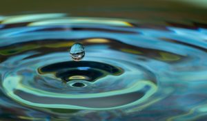 A close-up photograph of a single water droplet about to hit a pool of water; pool of water is causing a circular ripple motion. Water, in this case, signifies sustainability.