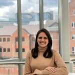 The Manager, Corporate Strategy & Development, ASDA and speaker for BGA Careers Month smiling and learning on a white table. She has medium length brown hair and brown eyes, wearing a loose light tanned colour polo neck long sleeve. She is standing behind a glassed window overlooking some buildings.