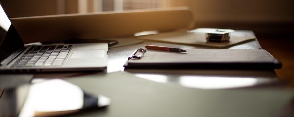 Dark office desk with a laptop, scattered papers, clip board and pen.