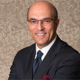 The Professor of Management, Dean of the School of Business at the American University in Cairo and speaker for AMBA & BGA Accreditation Forum, is grinning. The Dean wears clear oval glasses and is dressed in a smart white shirt, navy blue blazer, and a navy blue tie. The outfit is accompanied by a burgundy handkerchief emerging from the left pocket.